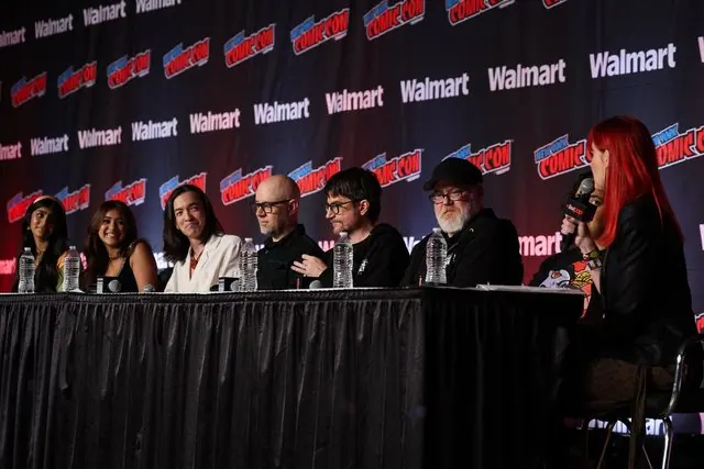 Angled view of the NYCC 2026 Avatar panel: the panelists at the table with the red-haired moderator (Janet Varney per the press release) holding a mic in the foreground